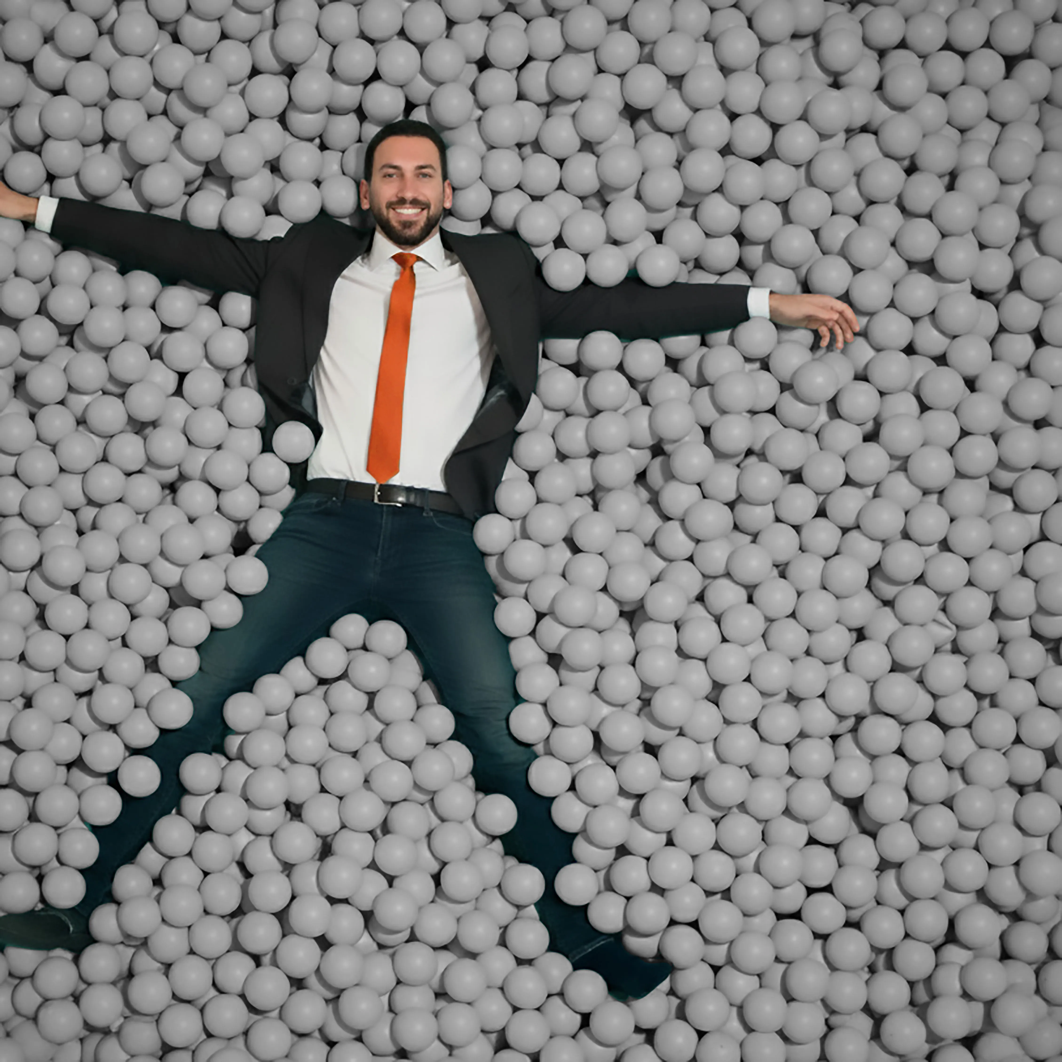Dan Limonchik in a suit, lying in a ball pit of grey balls, arms spread